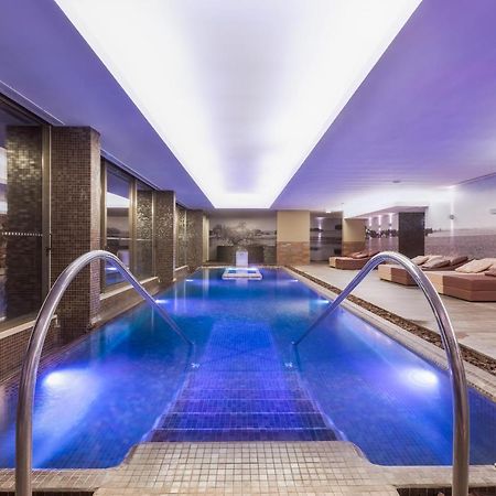 Ap Lago Montargil - Conference & Spa Exterior photo The photo shows a modern indoor swimming pool area. The pool features a sleek design with steps leading into the water and a gentle curve at the sides. The water is illuminated in a blue hue, enhancing the relaxing atmosphere. Surrounding the pool ar