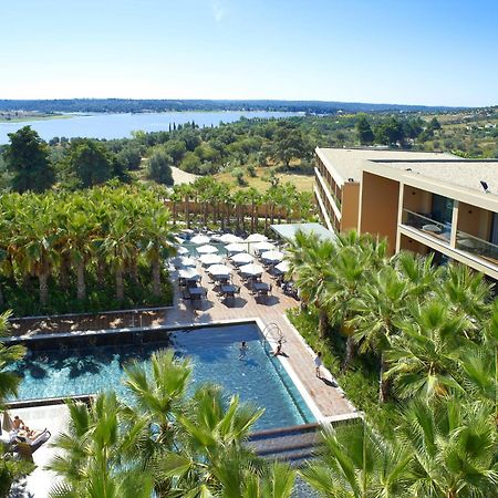 Ap Lago Montargil - Conference & Spa Exterior photo The photo shows a scenic view of a modern hotel or resort. In the foreground, there is a swimming pool surrounded by palm trees, with lounge chairs positioned beside it. Sun umbrellas provide shade over the chairs. The background features a lush land