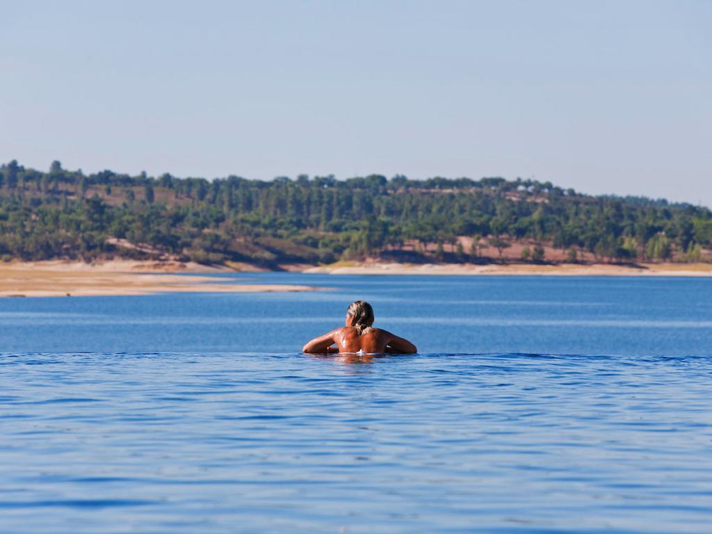Ap Lago Montargil - Conference & Spa Exterior photo The photo shows a person swimming in a calm body of water, likely a lake or a river. The swimmer is seen from behind and appears to be enjoying the water on a sunny day. In the background, there's a scenic landscape featuring green trees and a sandy 