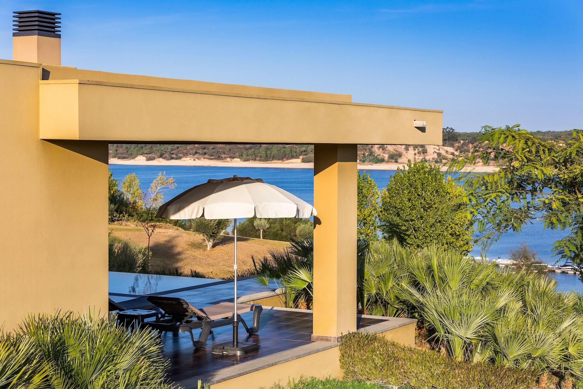 Ap Lago Montargil - Conference & Spa Exterior photo The photo shows a peaceful outdoor area with a modern architectural structure featuring an overhang. There is a lounge chair positioned beneath a large umbrella, creating a shaded spot to relax. In the background, there is a picturesque view of a bod