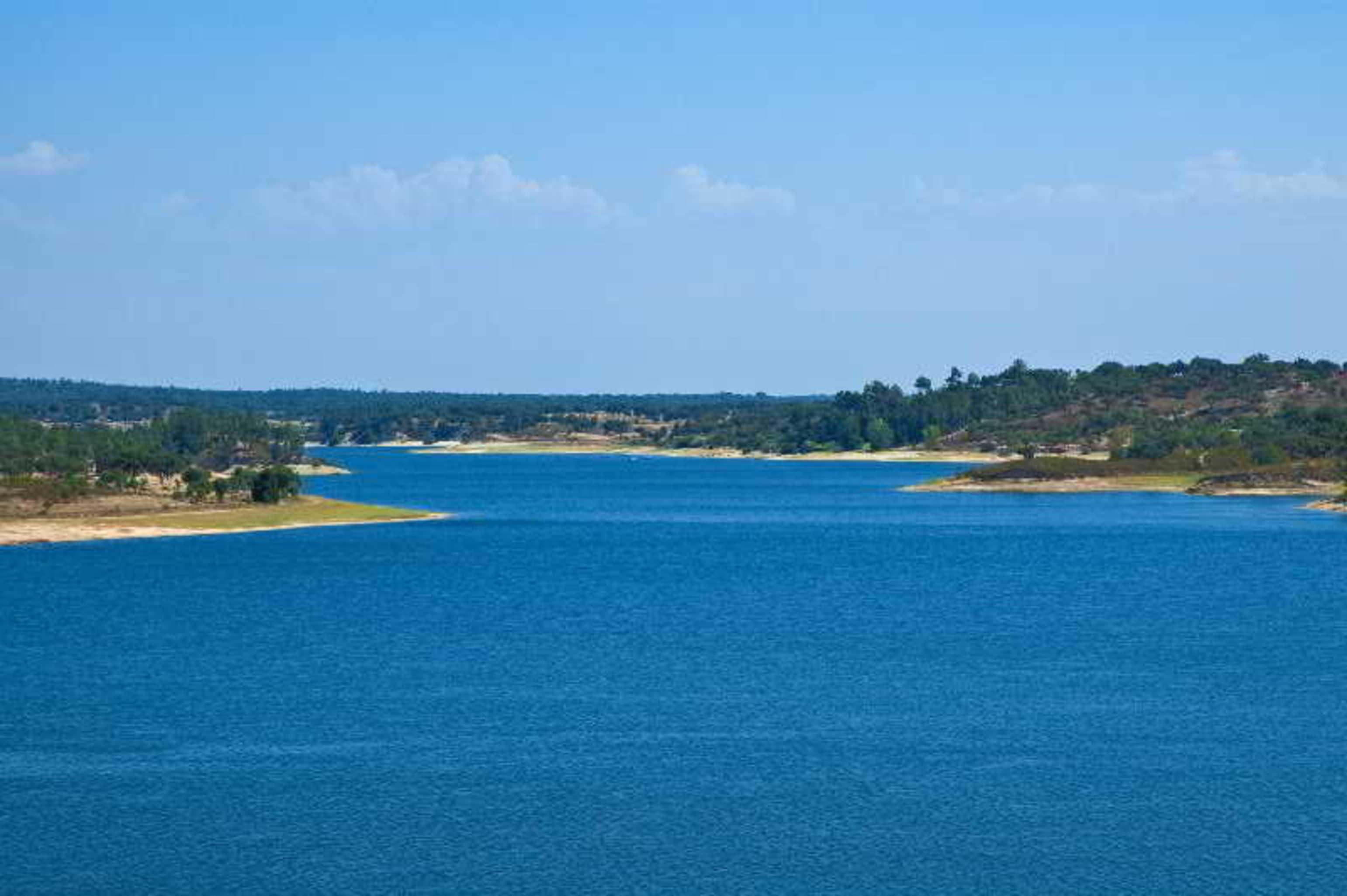 Ap Lago Montargil - Conference & Spa Exterior photo The photo depicts a serene landscape featuring a large body of water, likely a lake or river. The water appears calm and is reflecting a clear blue sky with some clouds. Surrounding the water, there are patches of green land and trees, indicating a n