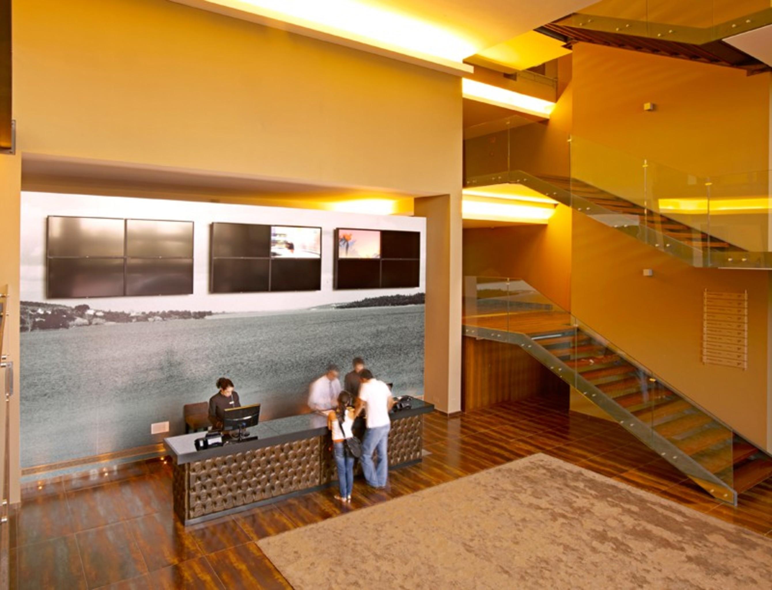 Ap Lago Montargil - Conference & Spa Exterior photo The photo depicts a modern lobby area with a reception desk. The design features a combination of warm colors and wood flooring. In the background, there is a large wall-mounted screen or display, showcasing several monitors. The wall behind the desk